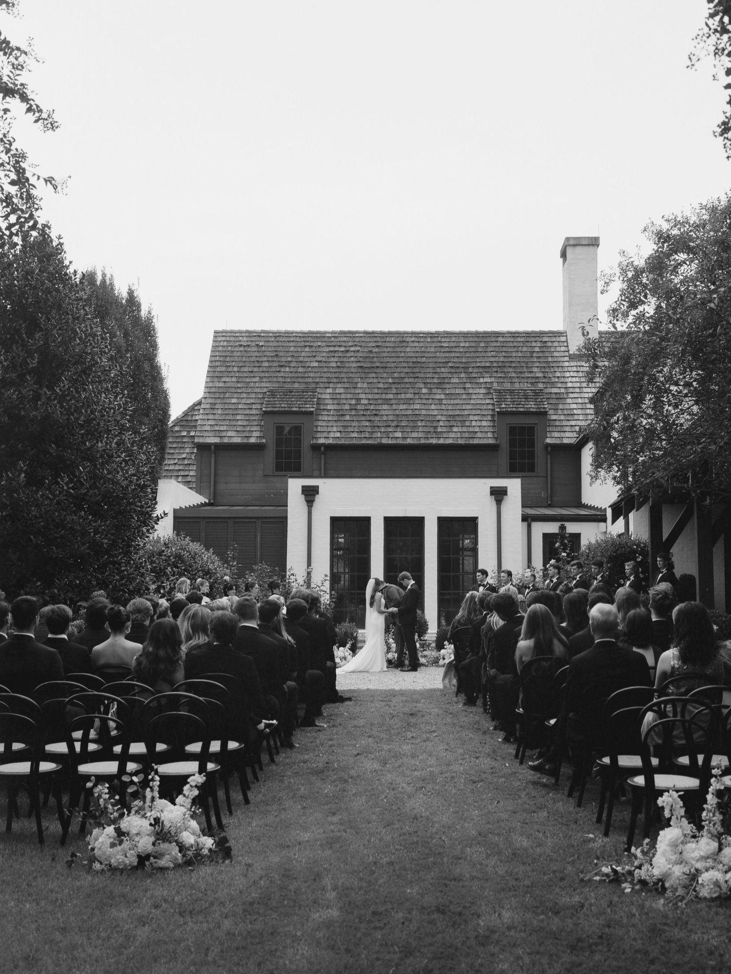 An outdoor wedding ceremony setup with rows of chairs facing a small white building surrounded by trees and greenery.