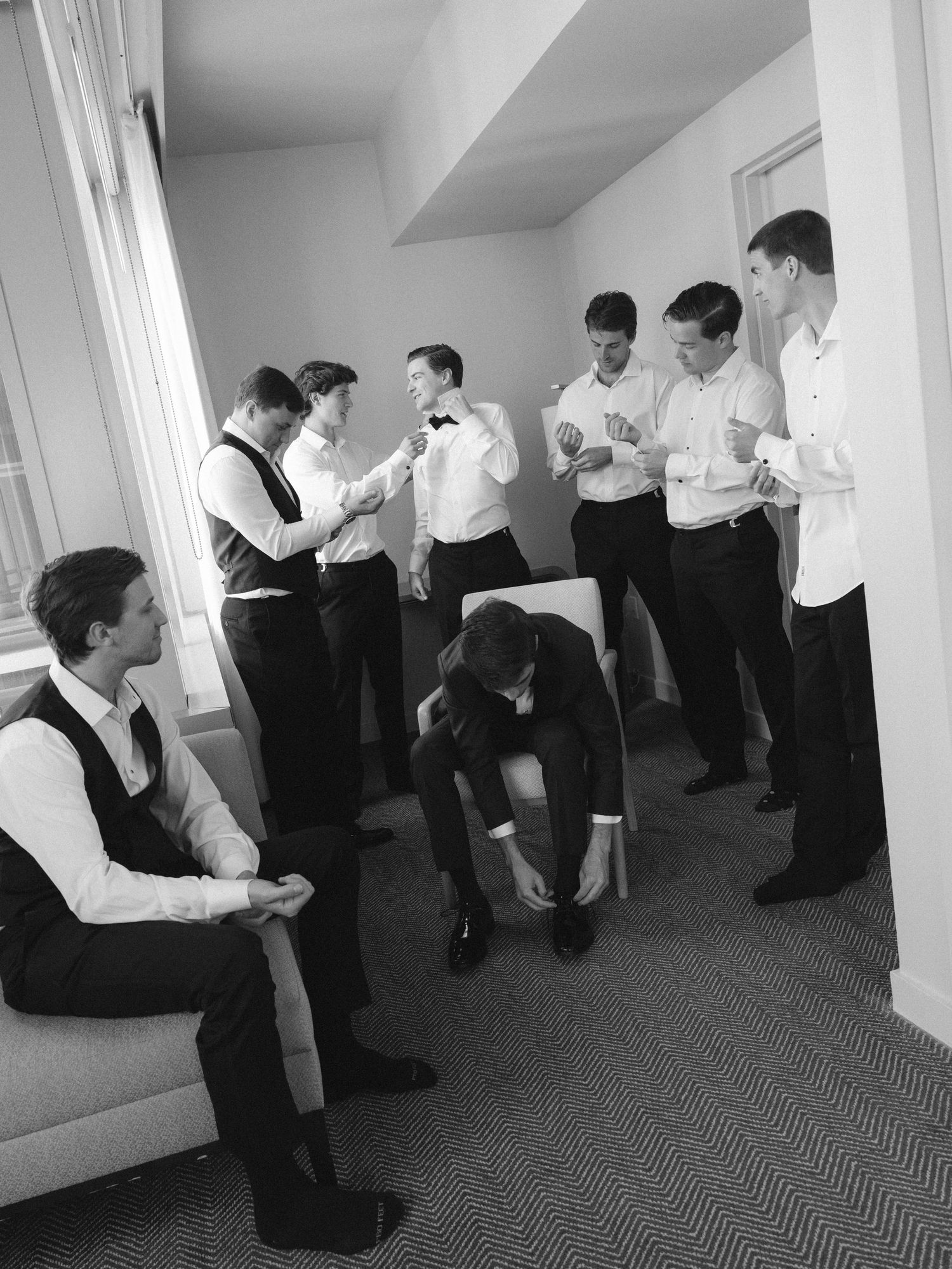Groomsmen in formal attire help each other prepare in a hotel room before a wedding ceremony.