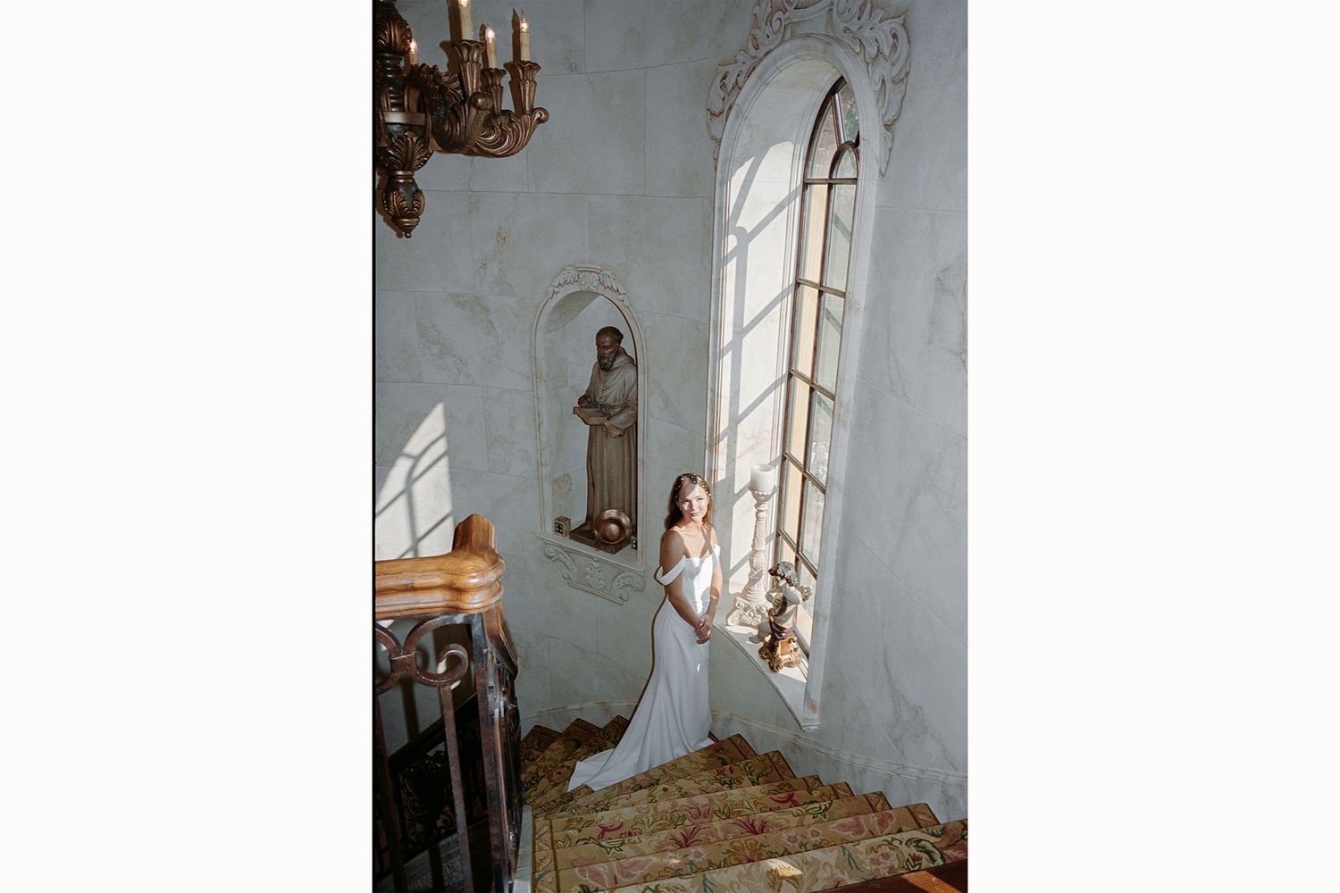 A person in a white dress poses gracefully against a curved stone staircase in an elegant interior setting.