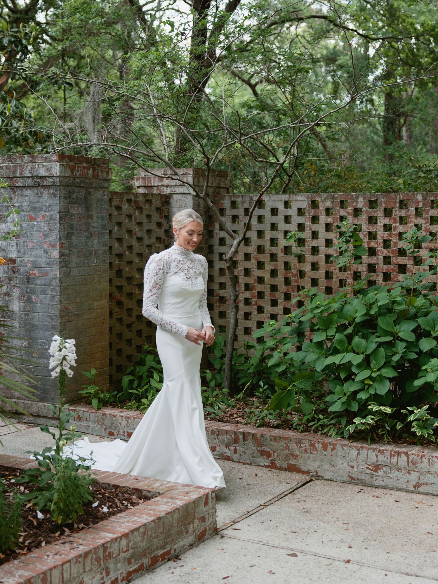 Bride in long-sleeve lace wedding gown standing in a lush garden with brick walls and greenery.
