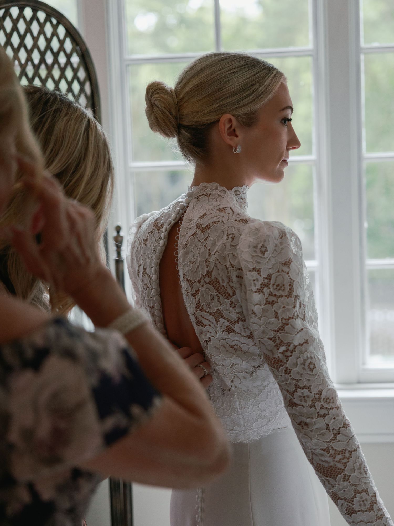 Bride in elegant white lace long-sleeve wedding dress with open back, standing by window during bridal preparations.