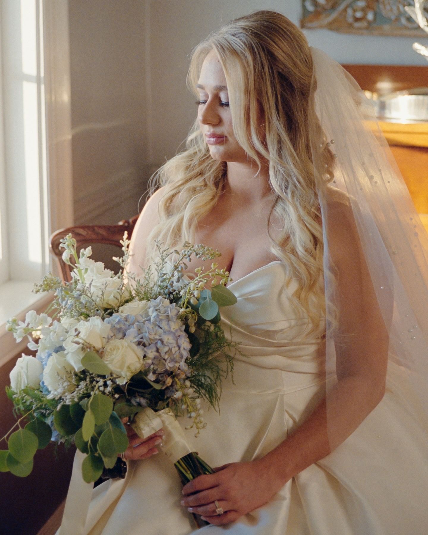 Elegant bride in white dress holding romantic bouquet of mixed flowers by window.