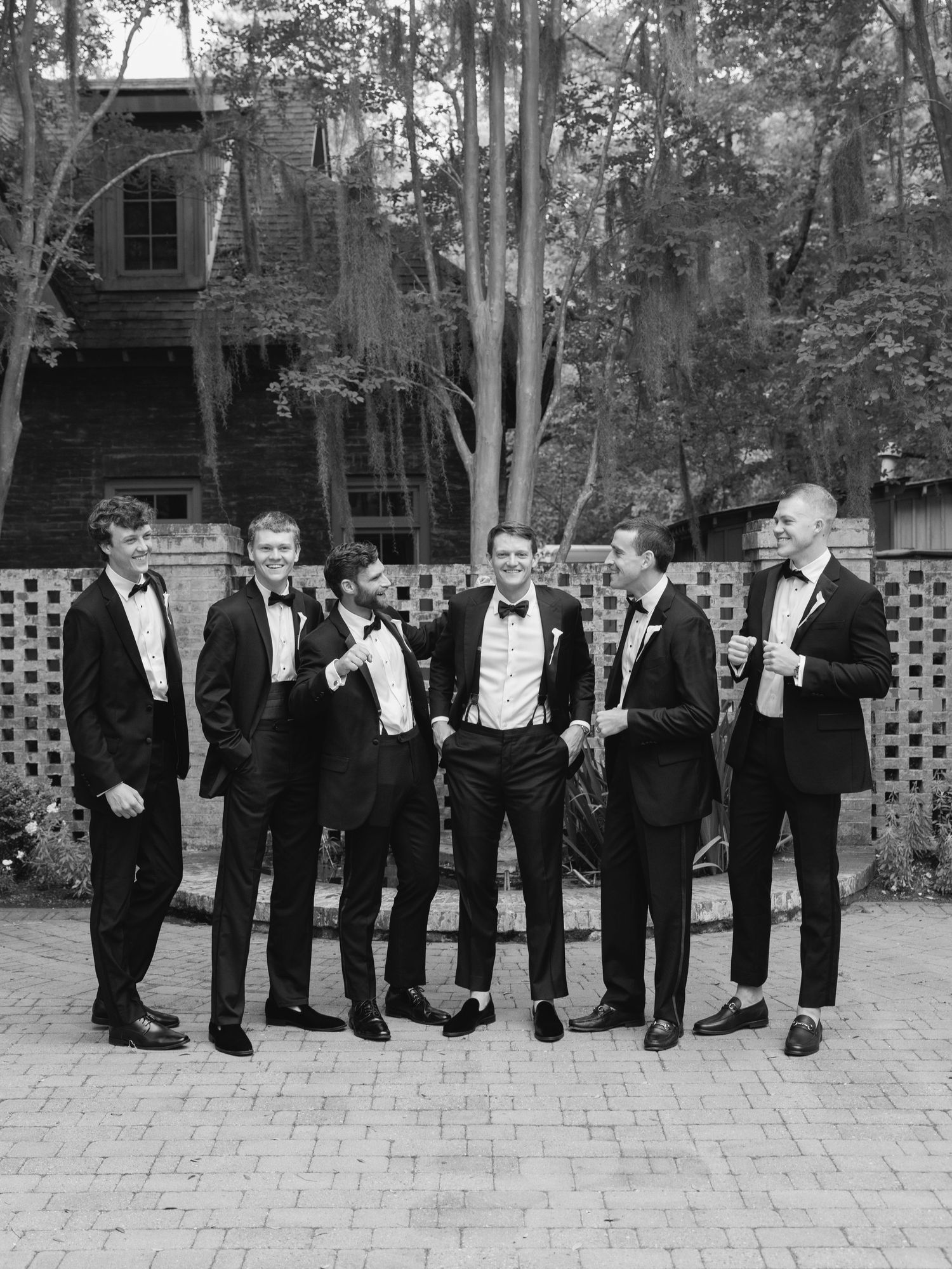 Groom poses with groomsmen in black tuxedos outdoors at elegant wedding venue.