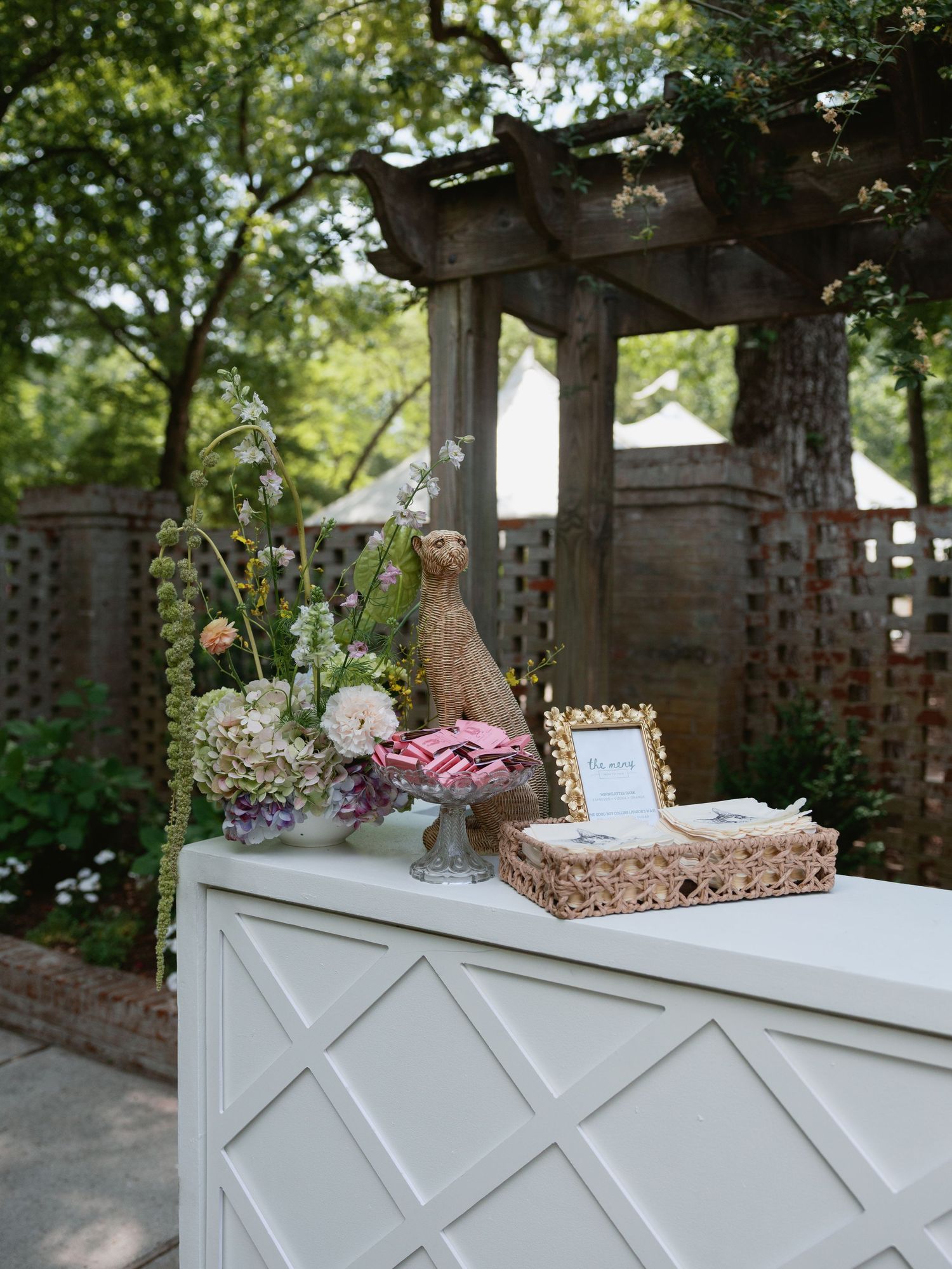 Elegant outdoor wedding welcome table with floral arrangement, decorative bunny figurine, and framed sign beneath a rustic wooden pergola.