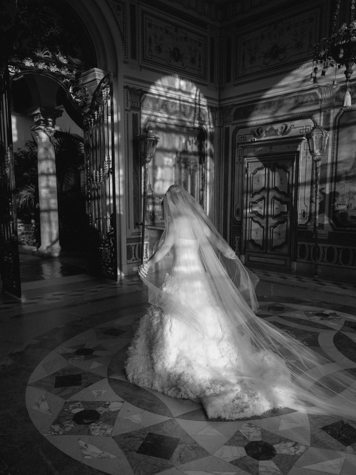 A bride in a flowing white wedding gown poses dramatically in a grand marble hallway with ornate architectural details.