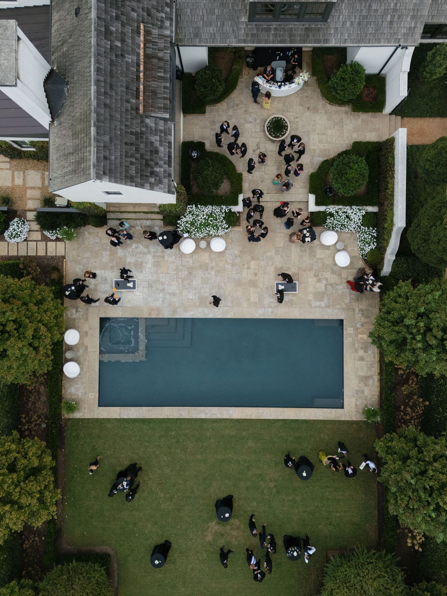 Aerial view of an outdoor event space with guests seated in black chairs on a lawn next to a rectangular pool.