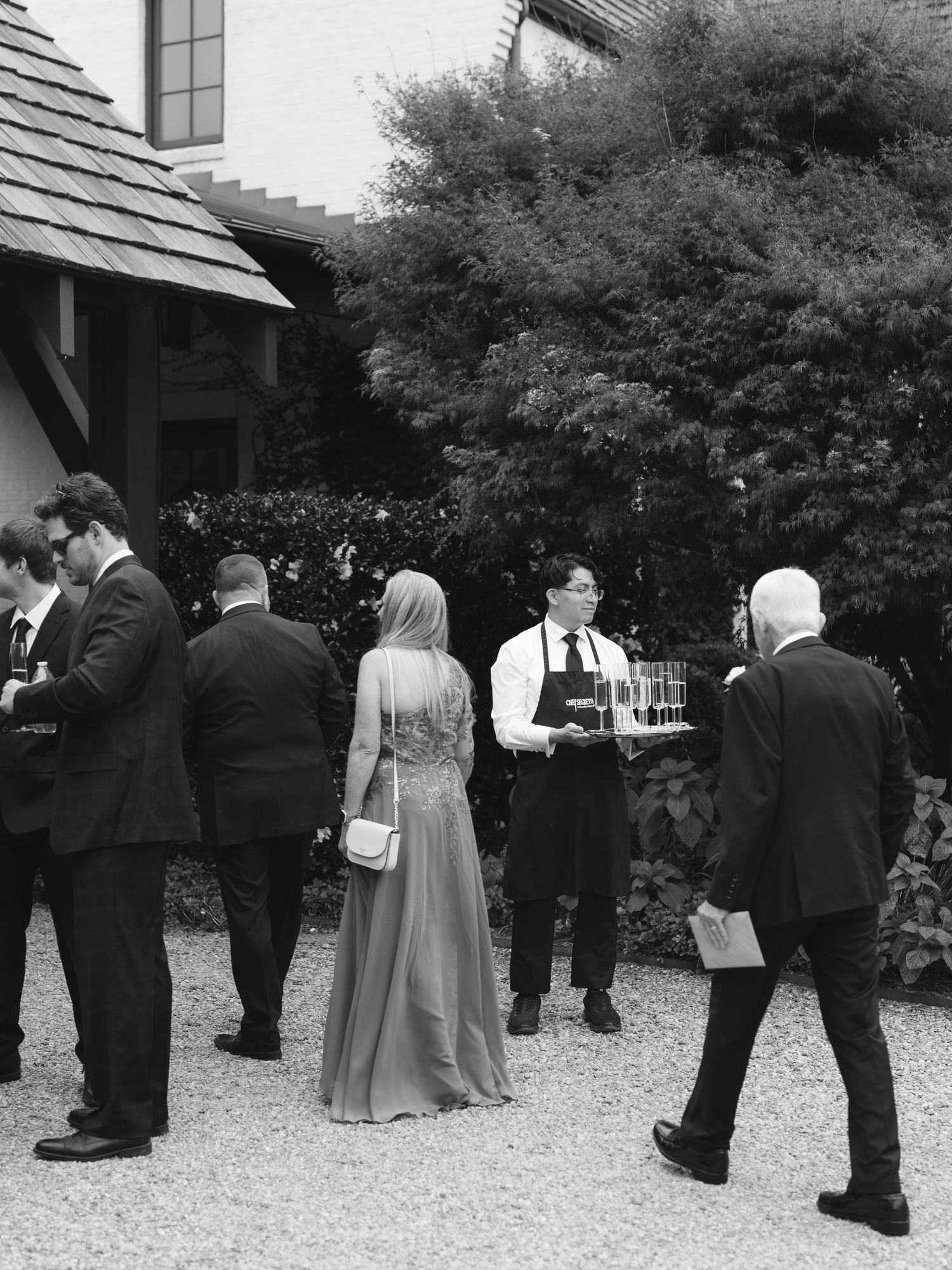 Black and white photo of guests mingling at an outdoor event with servers offering drinks on a gravel path.