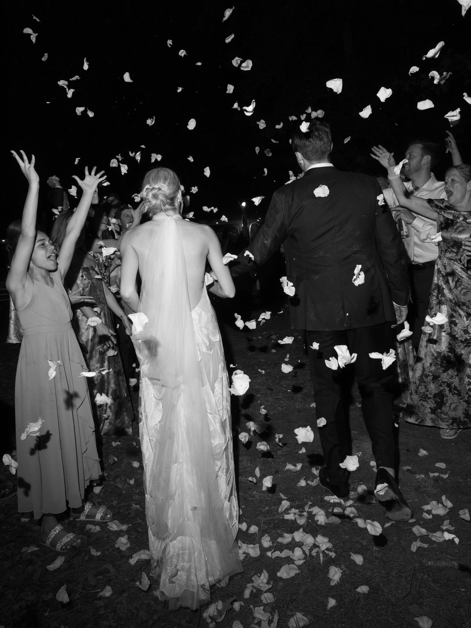 Black and white photo of bride and groom walking through flower petal toss at outdoor wedding celebration.