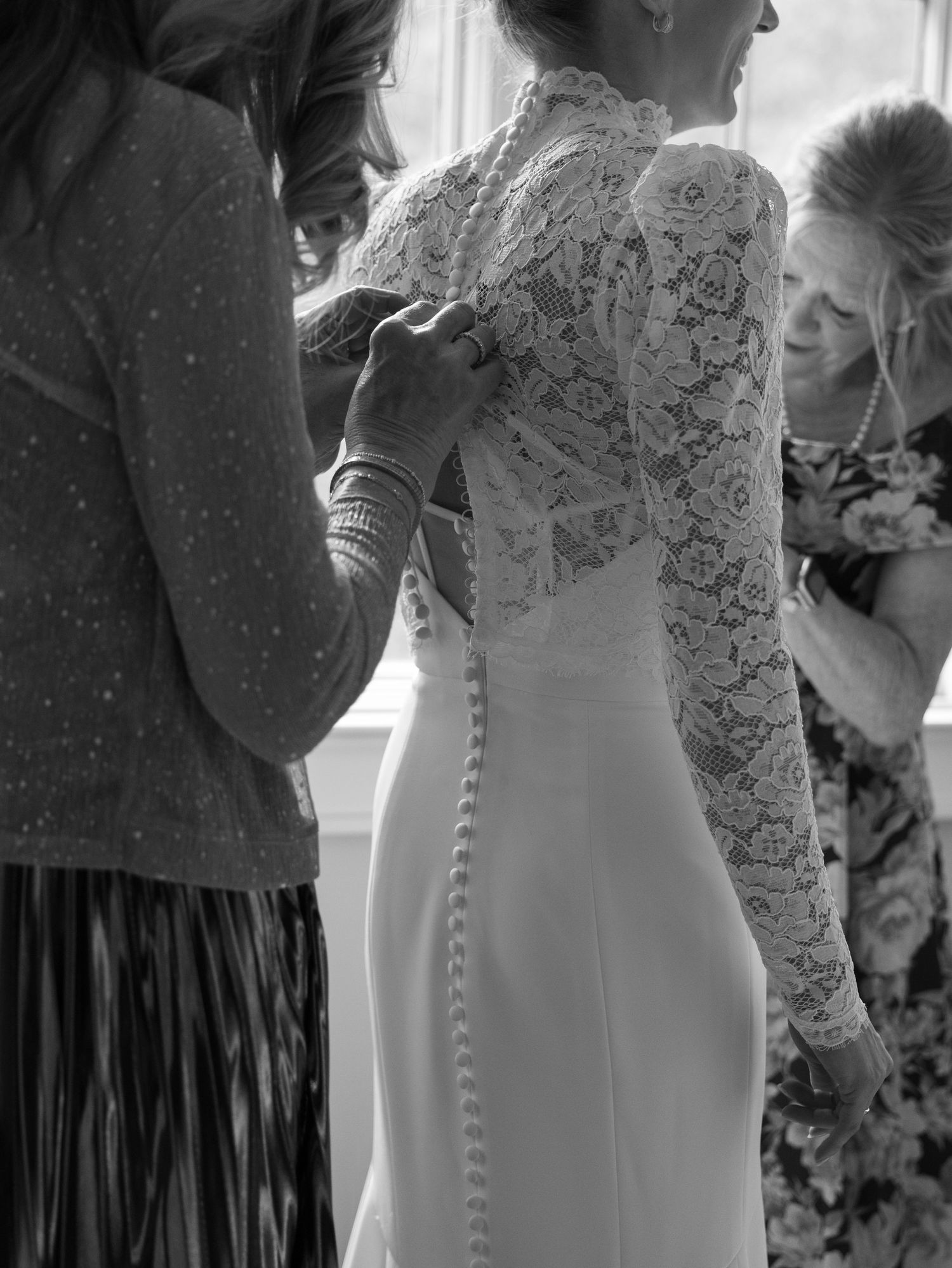 Black and white photo of bridesmaids buttoning the back of a lace wedding gown with pearl buttons.