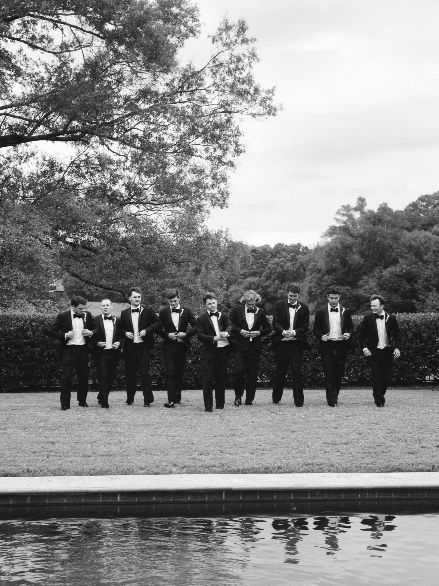 A row of formally dressed people in black tuxedos stand in a line along a reflecting pool in an outdoor garden setting.