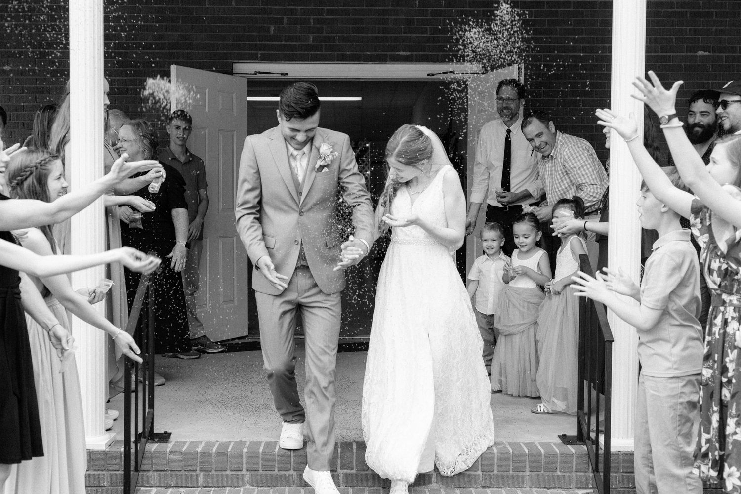 A black and white wedding celebration scene shows a couple exiting a building as guests cheer with extended arms.