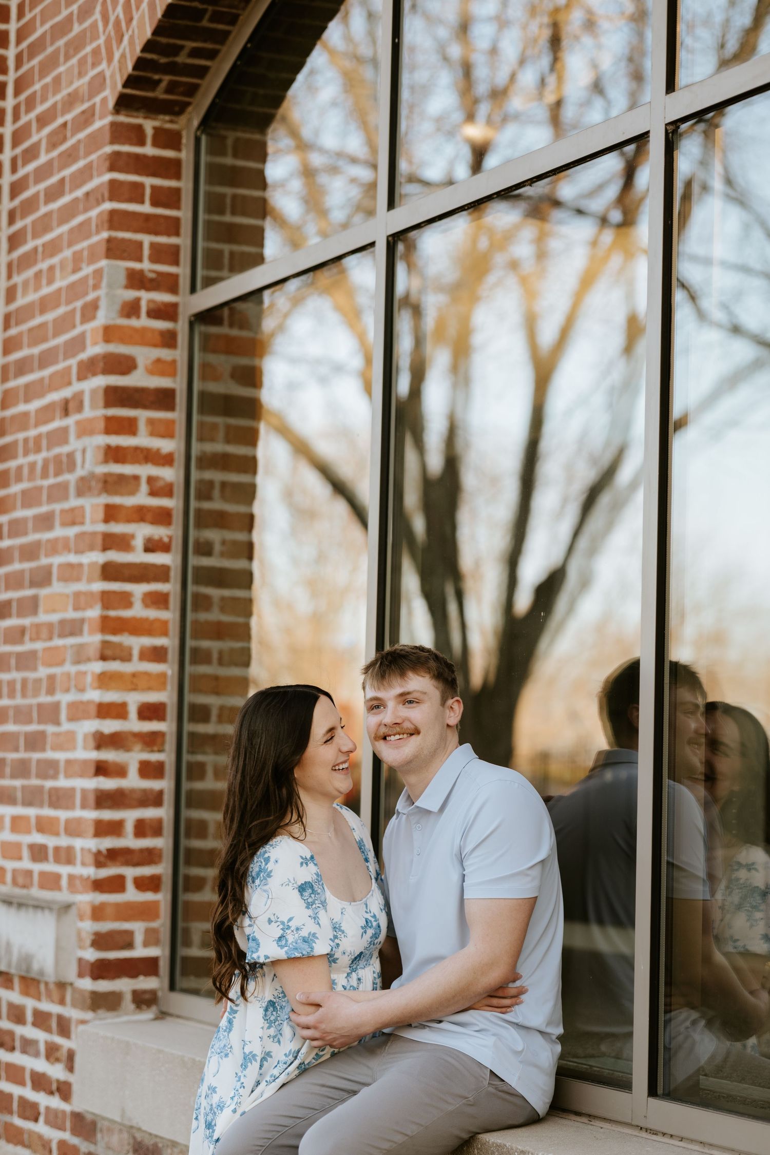A couple shares a tender moment while sitting outside a brick building with large windows at sunset.