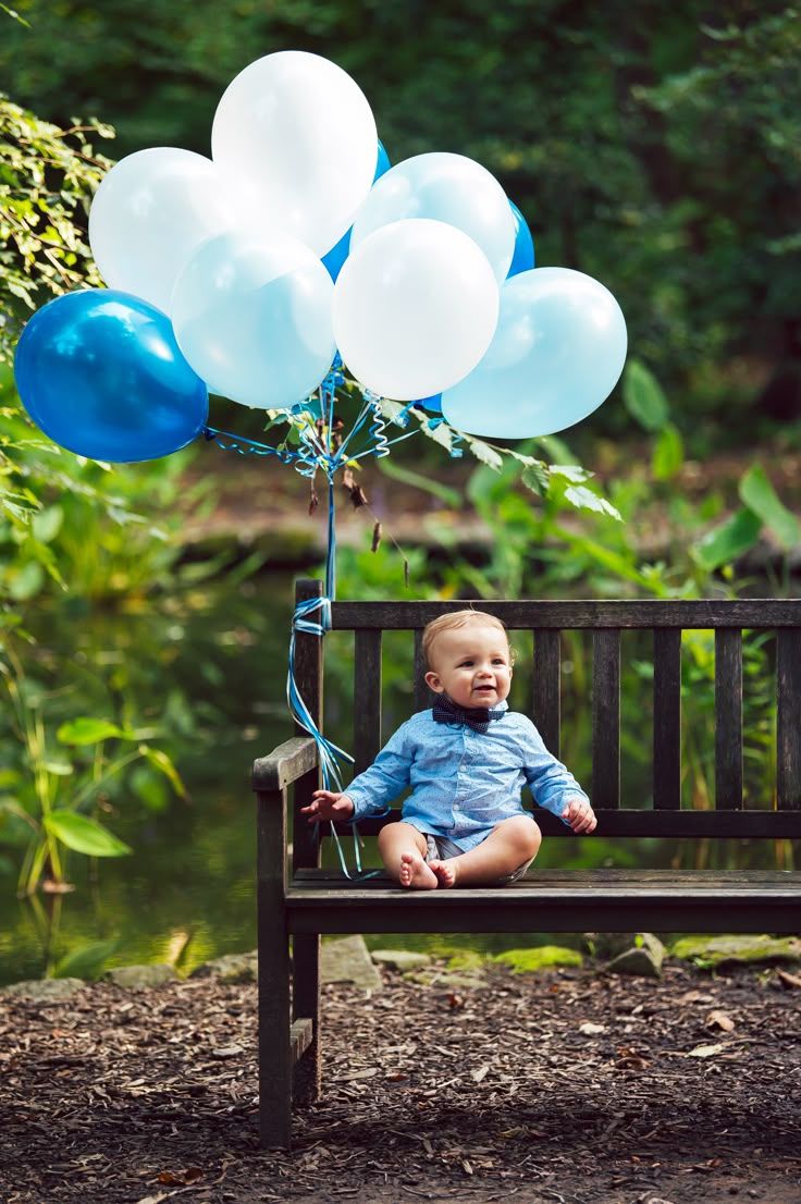 Baby boy in blue shirt sitting on park bench with blue and white balloon bouquet outdoors.
