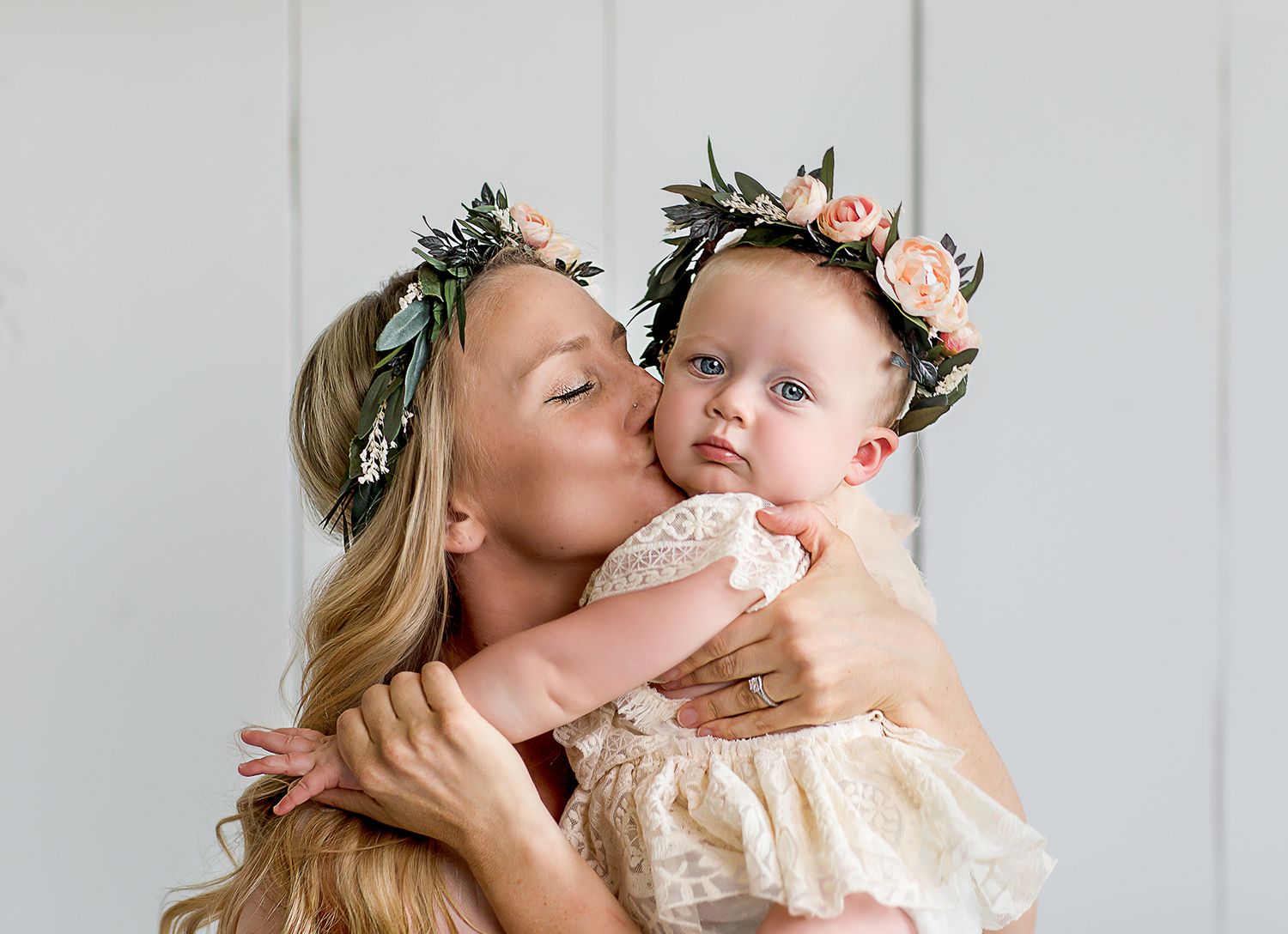 Blonde mother kissing baby girl both wearing floral crowns in soft white studio setting.