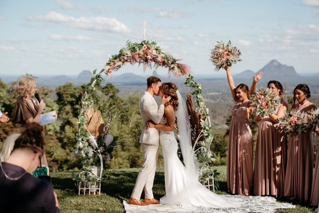Couple kissing under a floral arch with bridesmaids in mauve dresses at a scenic hilltop wedding ceremony.