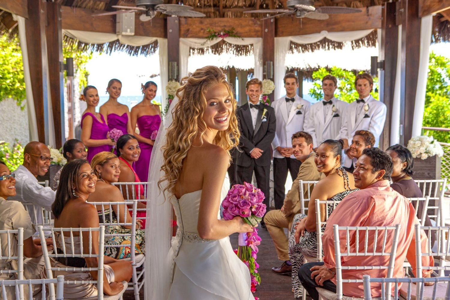 Bride in white gown holding purple bouquet walks down aisle at outdoor wedding ceremony with guests and bridal party.