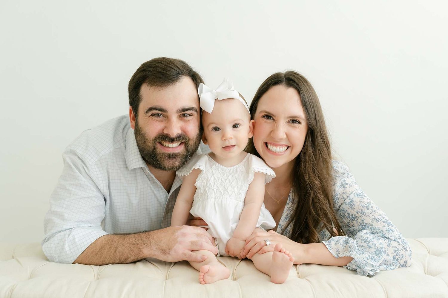 Smiling parents lying down with baby girl in white bow headband in bright studio portrait.