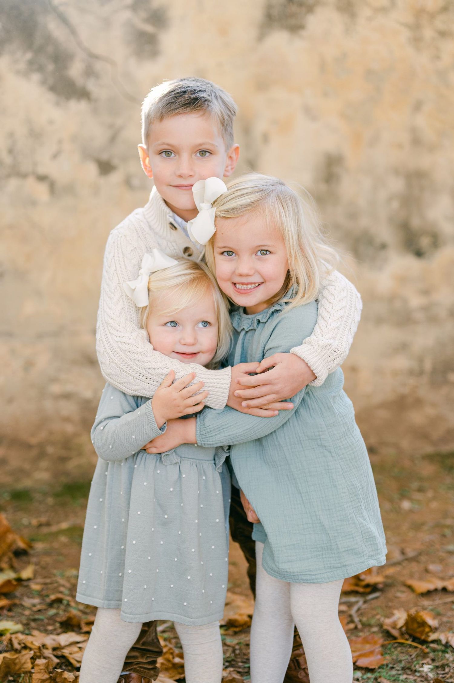 Three blonde siblings hugging each other warmly in front of a rustic textured wall in autumn.