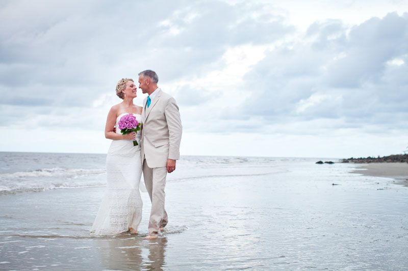 Bride and groom walking on beach shoreline, bride holding pink bouquet, groom in tan suit, ocean waves behind them.