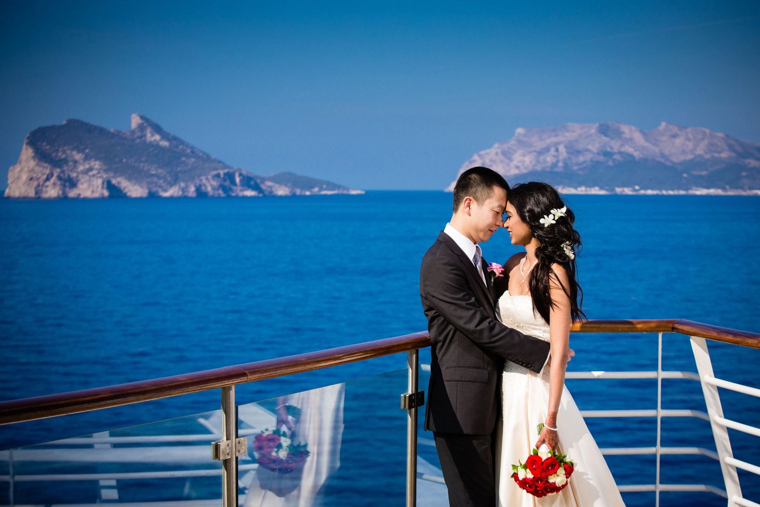 Newlywed couple sharing a romantic kiss on a cruise ship deck with stunning blue ocean and rocky islands backdrop.