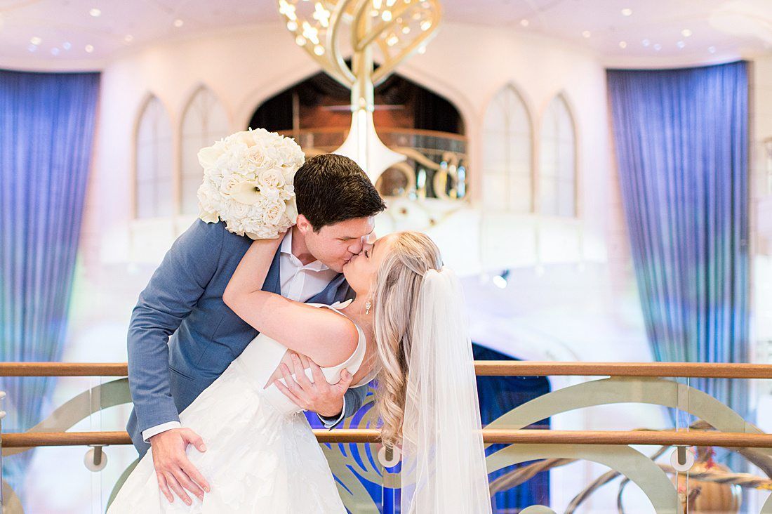 A bride and groom share a romantic kiss in an elegant blue and gold wedding venue with a chandelier backdrop.