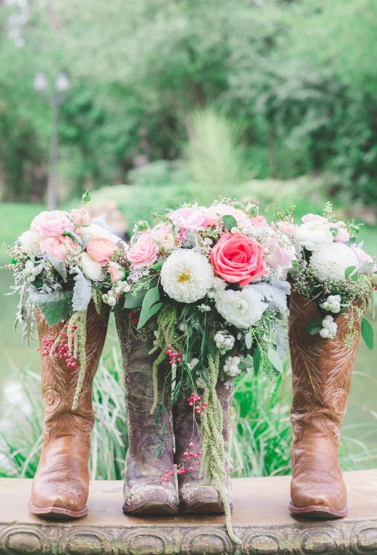 Three brown cowboy boots used as vases holding pink and white floral bouquets with greenery, displayed outdoors near a pond.