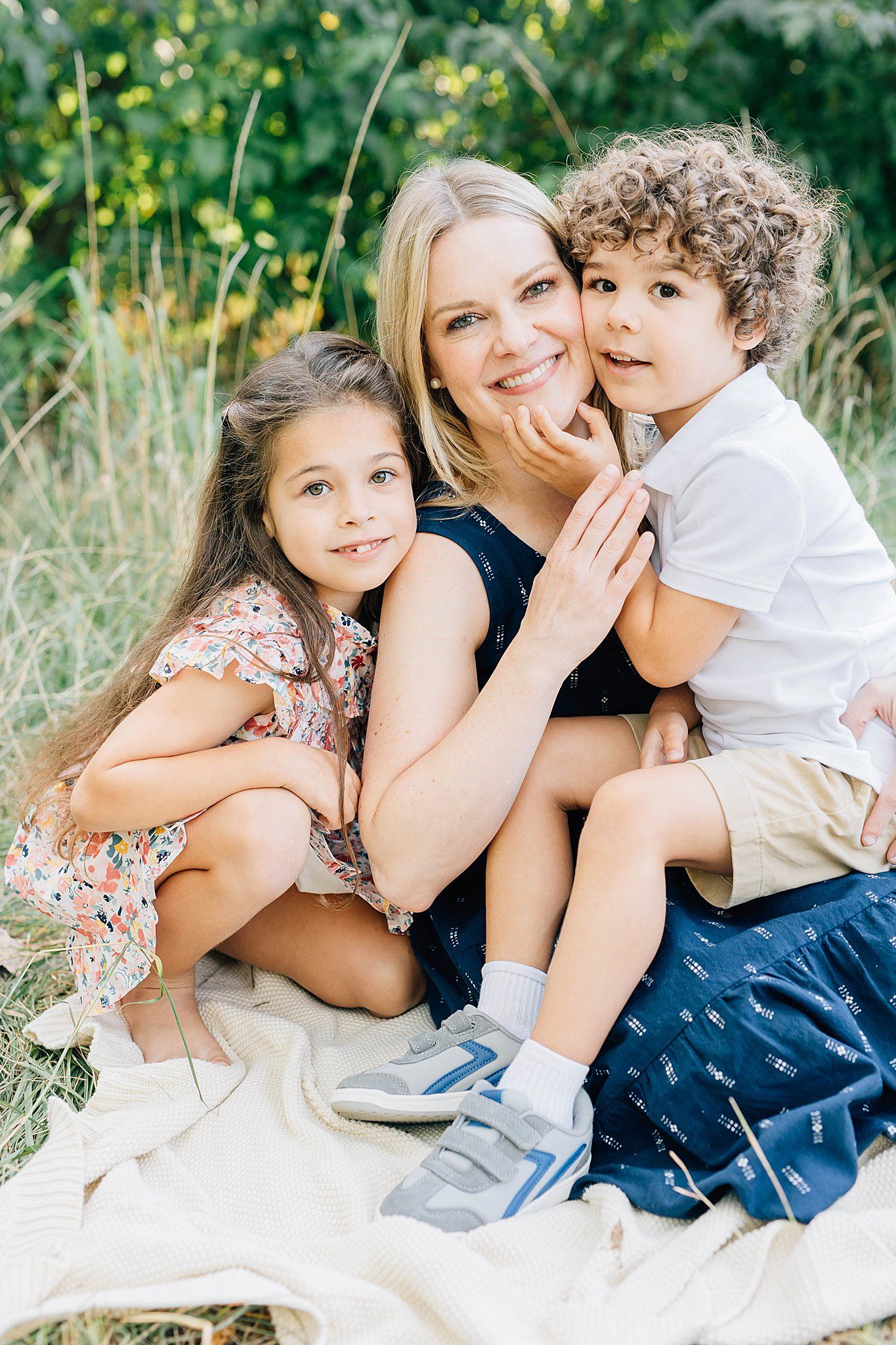 Smiling blonde mother sitting on a blanket outdoors with her two young children hugging her closely.