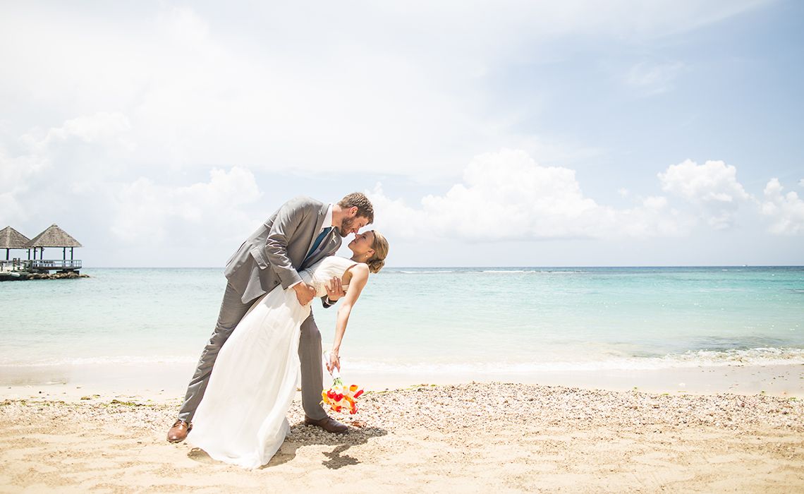Bride and groom share a romantic dip kiss on a tropical beach with turquoise ocean waters and overwater bungalows.