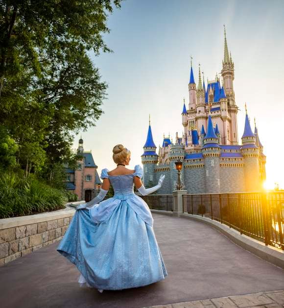 Cinderella in a blue gown walks toward a sunlit fairy tale castle at Disney World Magic Kingdom.