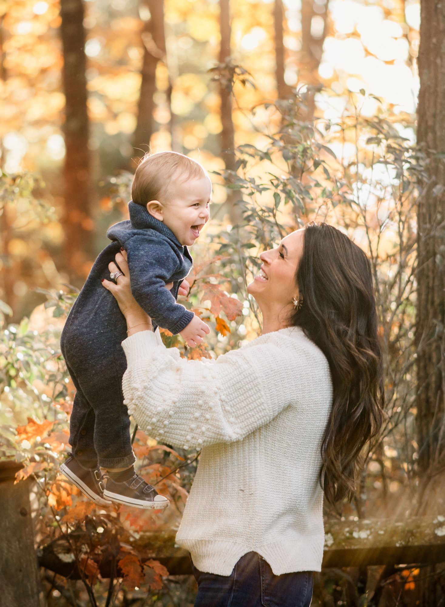 Mother lifting laughing baby in autumn forest with golden fall foliage in background.