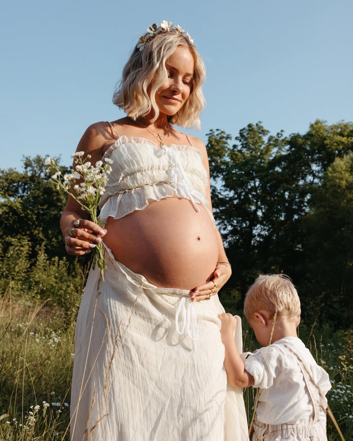 A pregnant woman in a white dress holds wildflowers while her toddler touches her bare baby bump.
