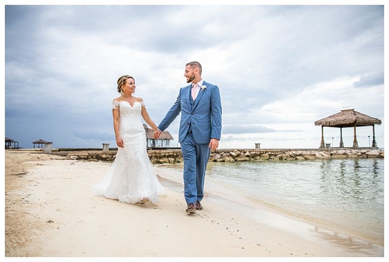 Bride and groom walk hand in hand on a tropical beach, with ocean views and a gazebo backdrop on their wedding day.