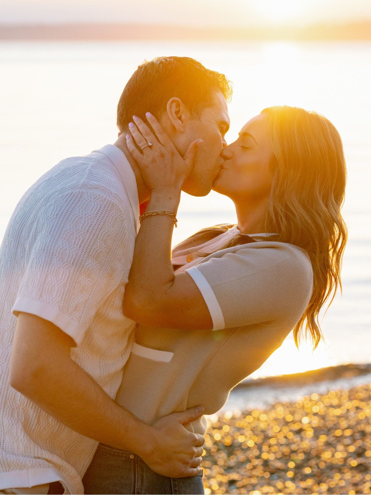 Couple sharing a passionate sunset kiss on a beach with golden light reflecting off the water behind them.