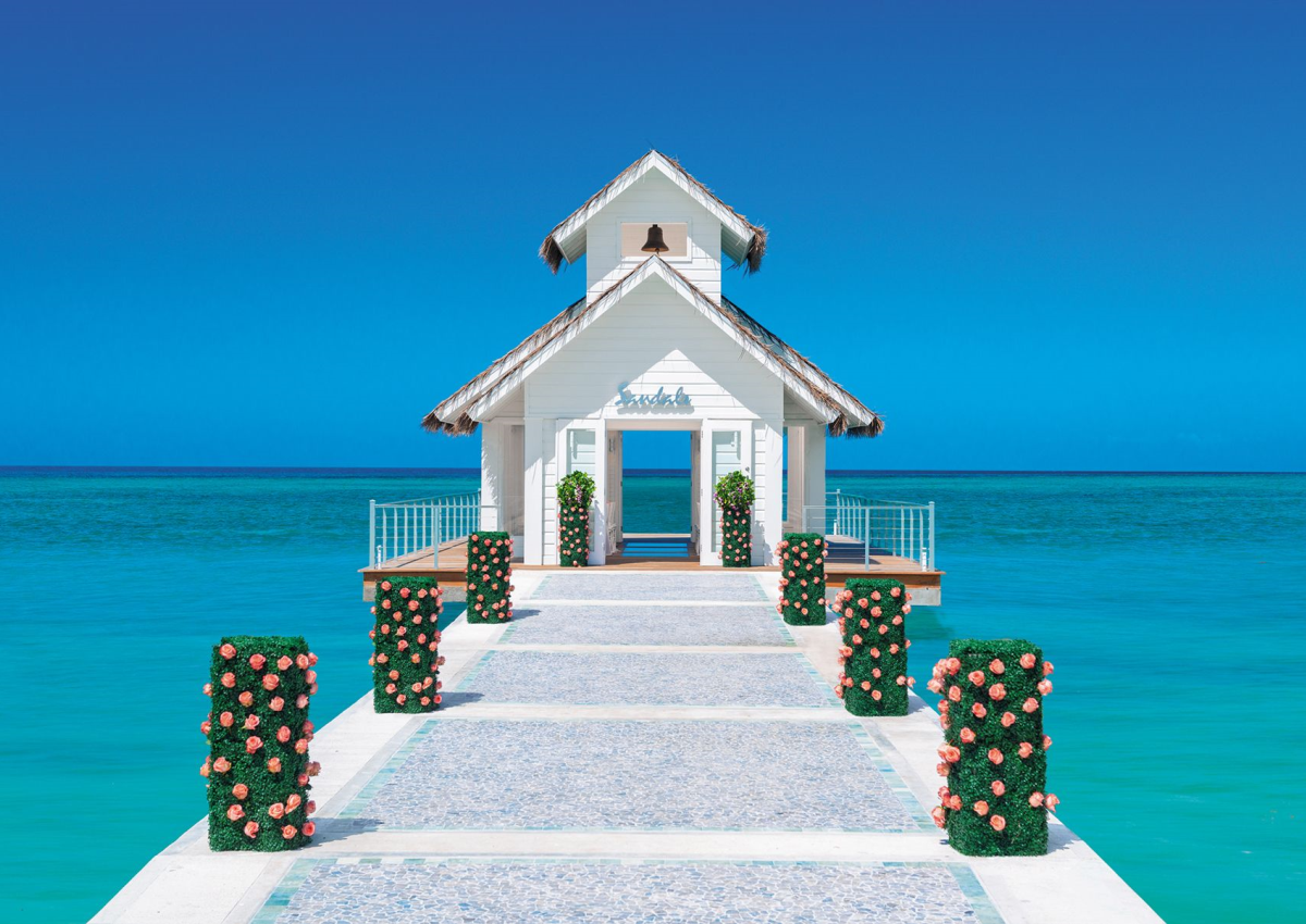 White Sandals chapel on a pier surrounded by turquoise Caribbean waters, adorned with pink roses under a bright blue sky.