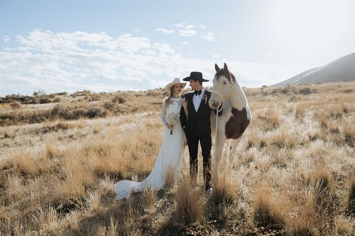 Bride and groom in western attire standing with a paint horse in a golden field under a bright blue sky.