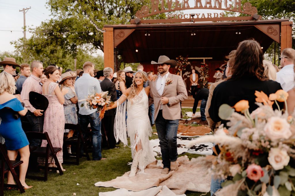 Bride and groom walk down the aisle at an outdoor rustic wedding ceremony with guests cheering.