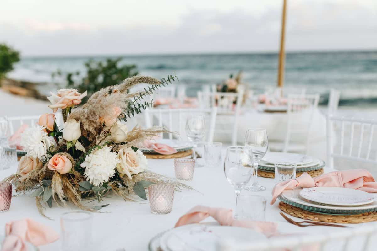 Elegant beach wedding reception table with blush roses, white hydrangeas, pampas grass centerpiece, and pink ribbon accents.