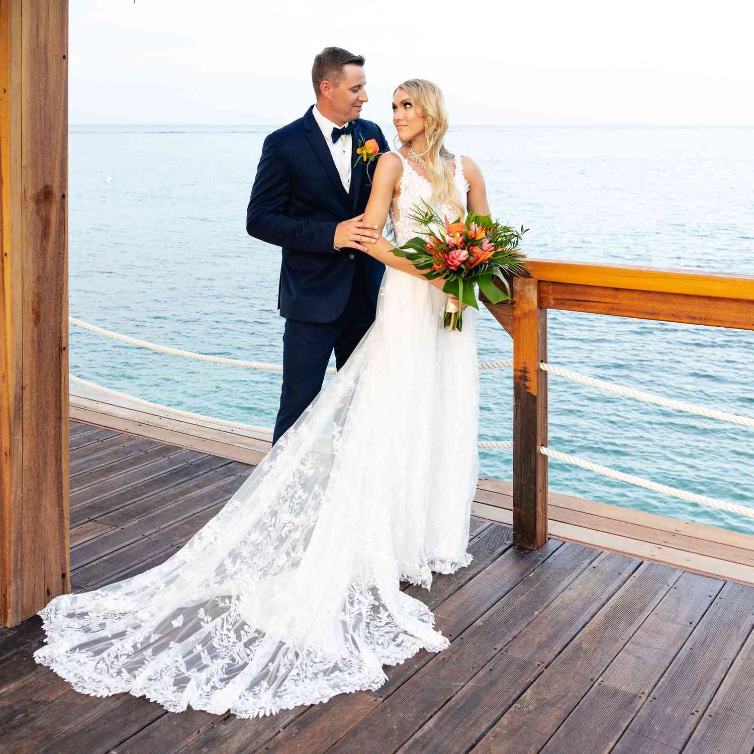 Bride in lace gown and groom in navy tuxedo pose on wooden deck overlooking turquoise ocean at tropical wedding.