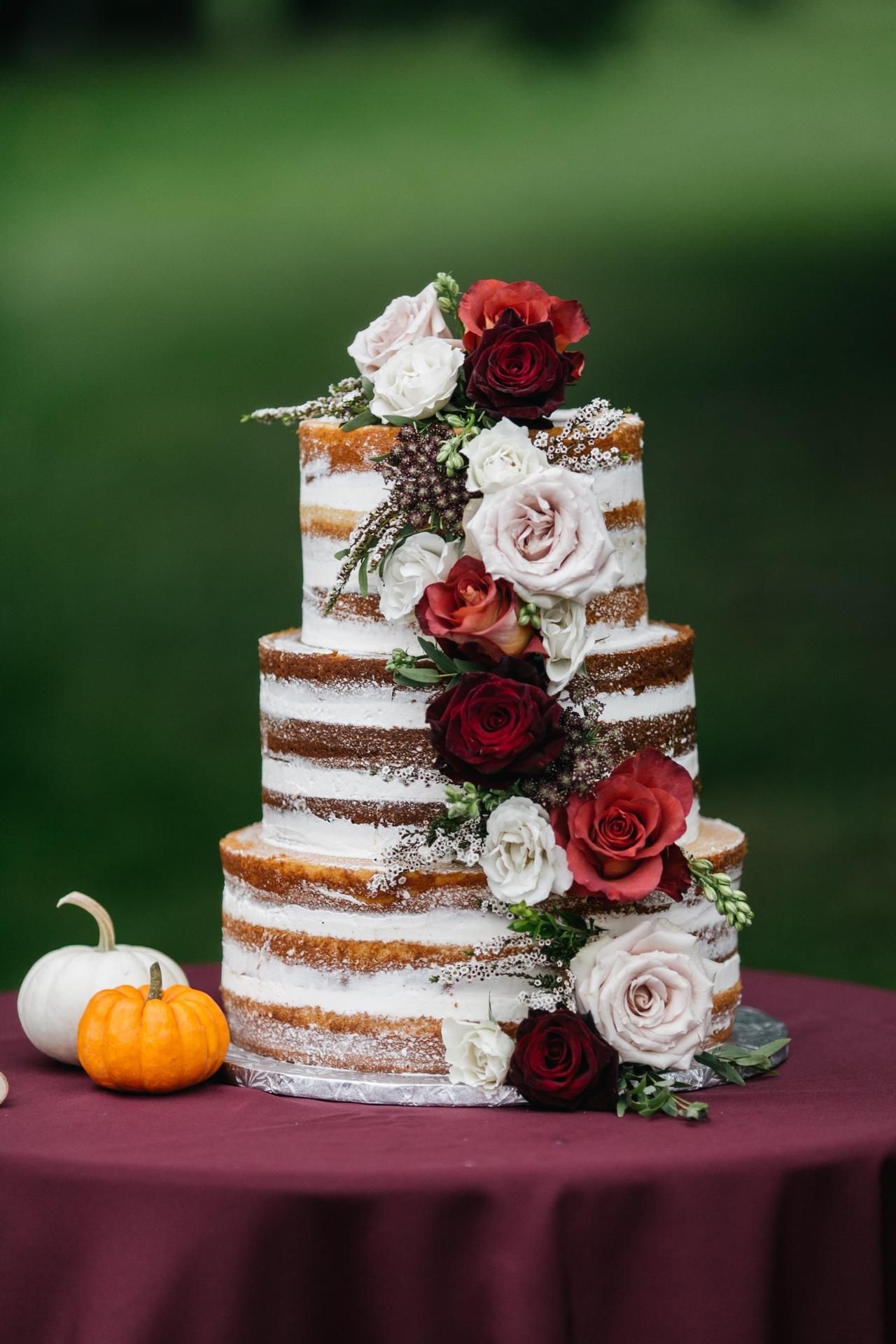 Three-tier naked wedding cake adorned with red and white roses on burgundy tablecloth with mini pumpkin.