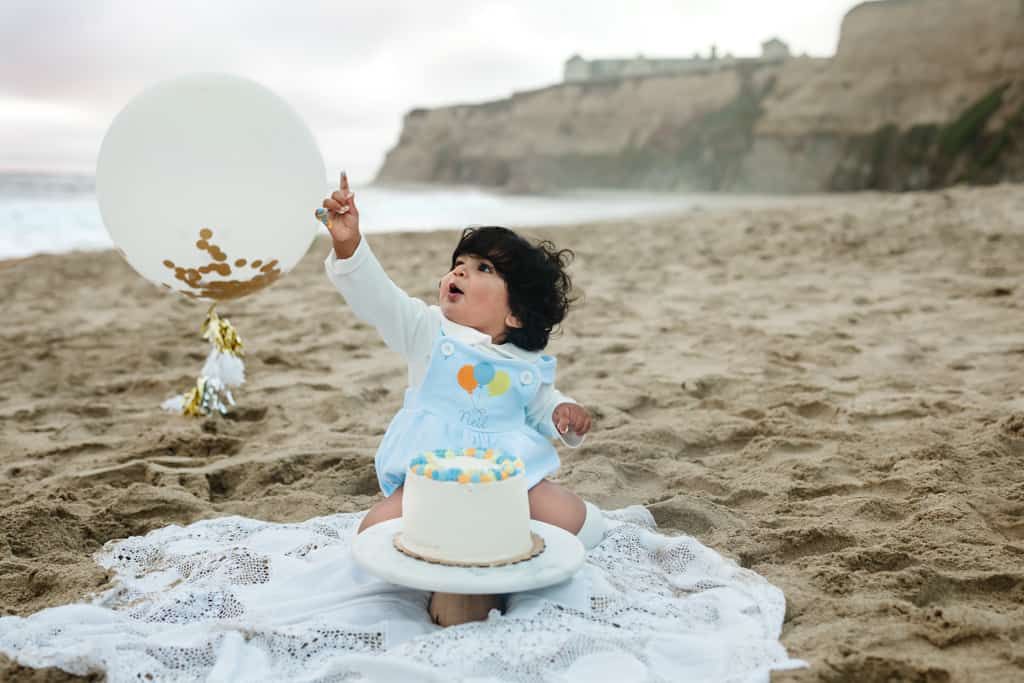 Baby's first birthday cake smash on beach with white balloon and decorated cake on blanket.