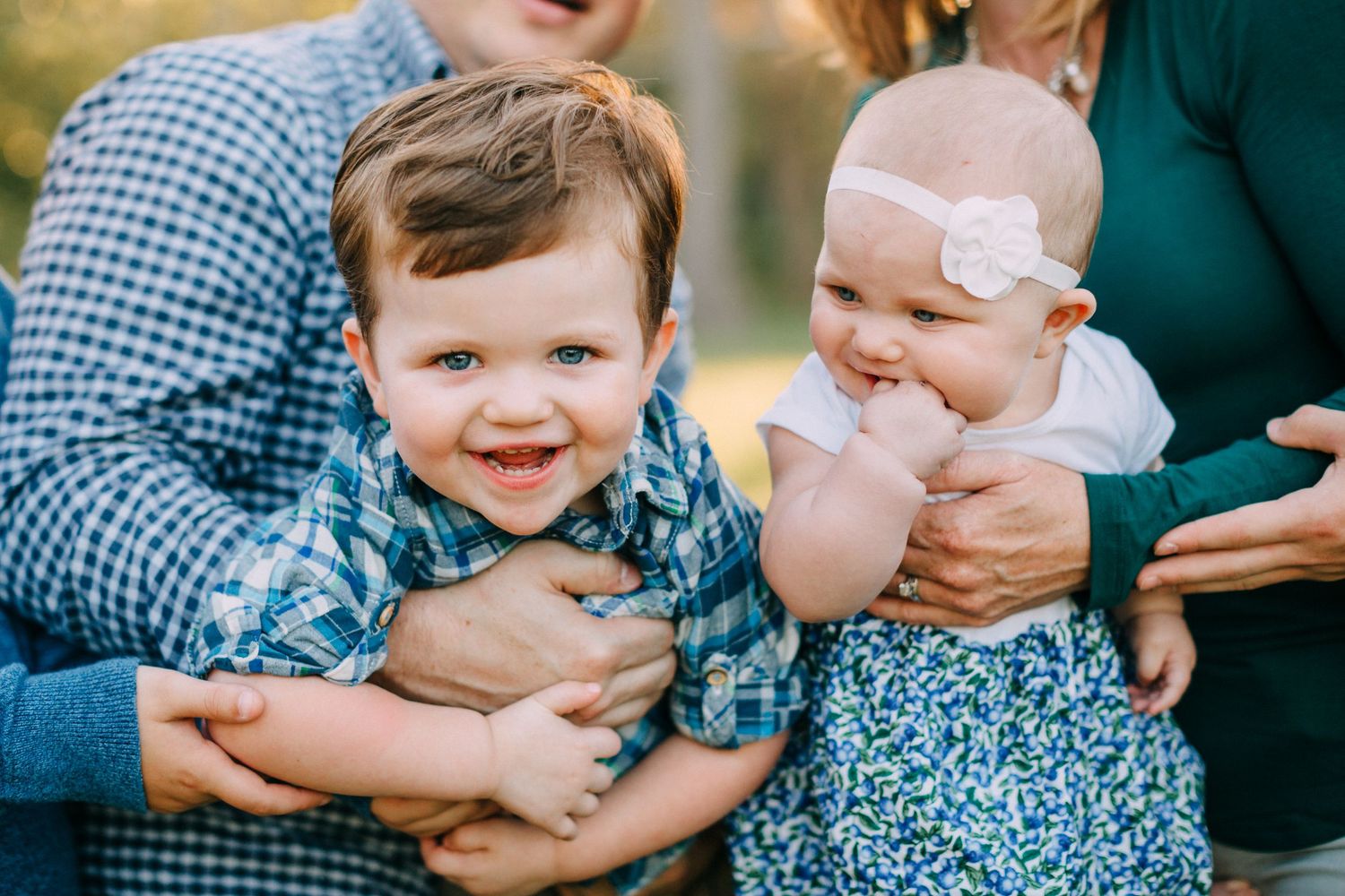 A smiling toddler boy and baby girl in floral outfit held by parents during an outdoor family photo session.