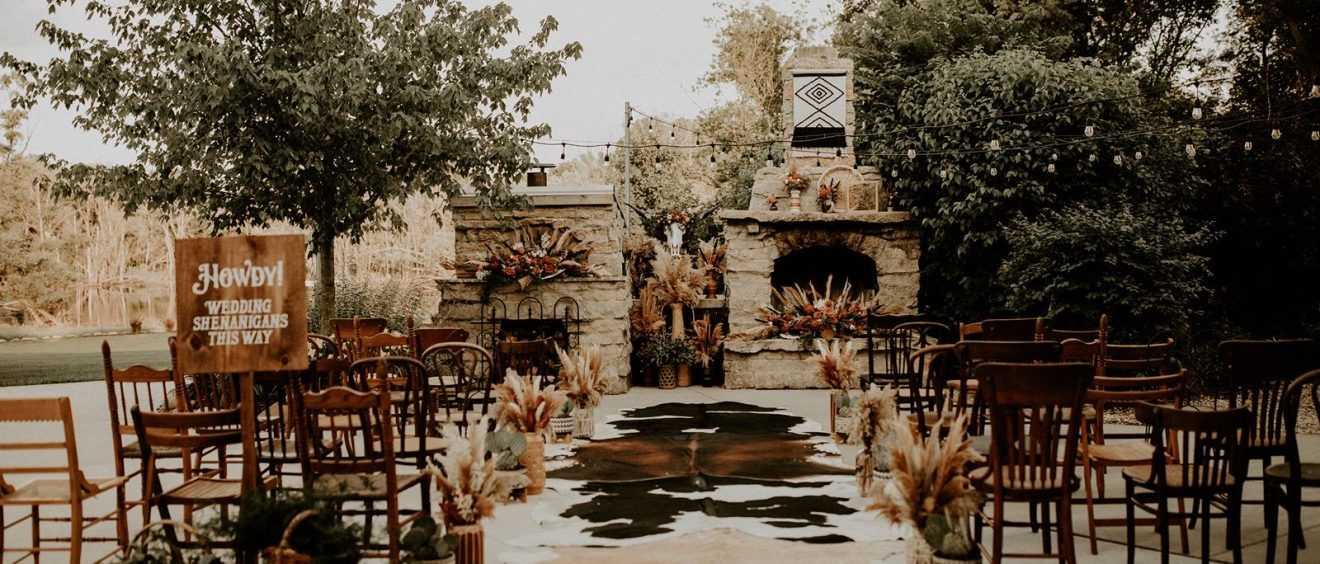Rustic outdoor wedding ceremony with wooden chairs, pampas grass, and a stone fireplace altar surrounded by lush greenery.