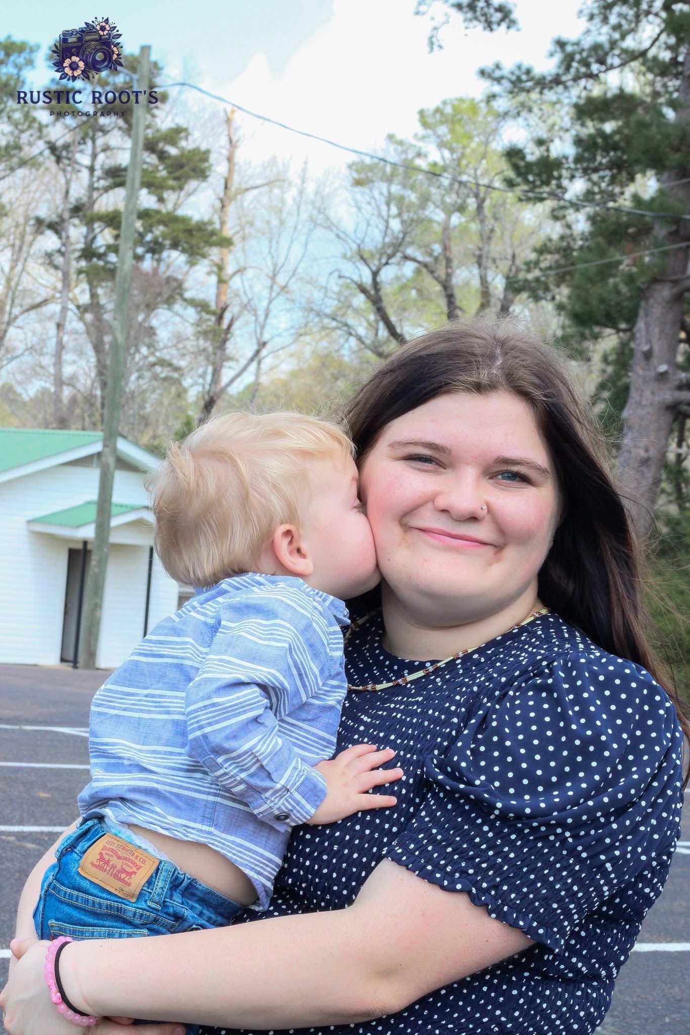 A young woman in a navy polka dot dress smiles while holding a baby boy who gives her a sweet kiss on the cheek.
