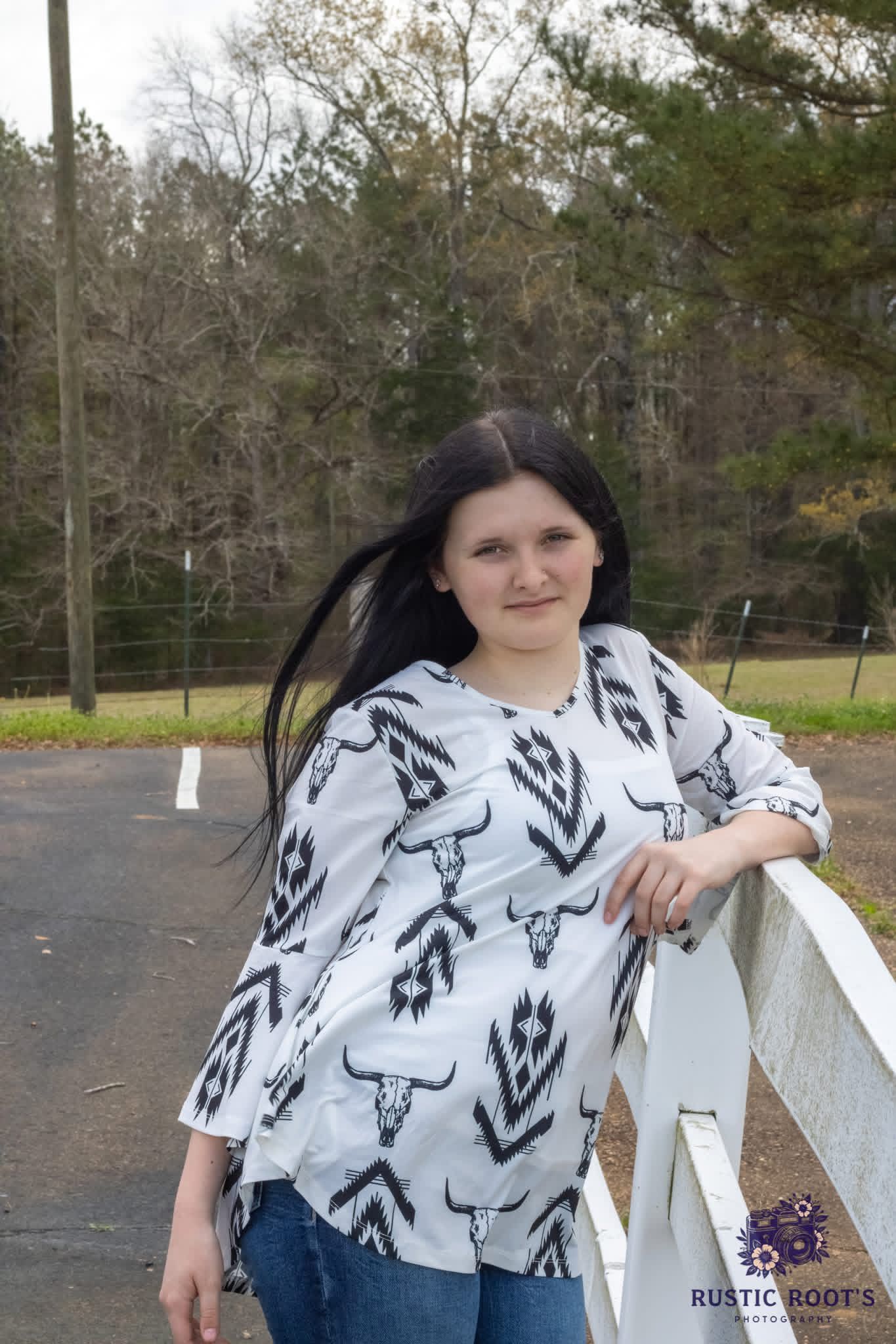 Woman in white aztec bull skull print top and jeans leaning on white fence with trees in background.