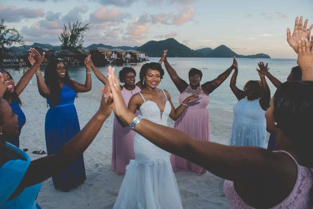 Bride in white gown celebrates on beach with bridesmaids in pink and blue dresses, raising hands joyfully.