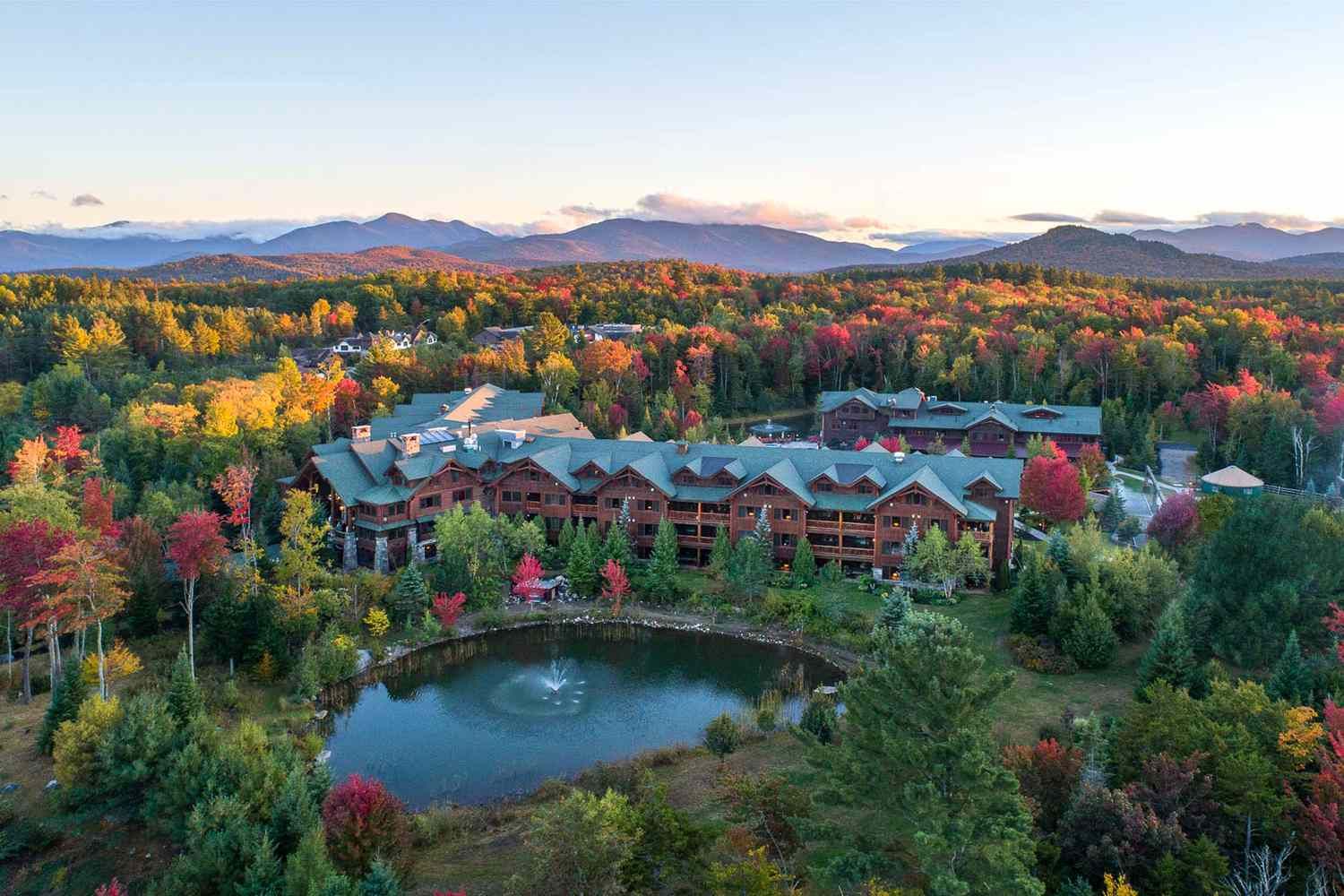Aerial view of a mountain resort surrounded by vibrant fall foliage, with a pond fountain and misty peaks at dusk.
