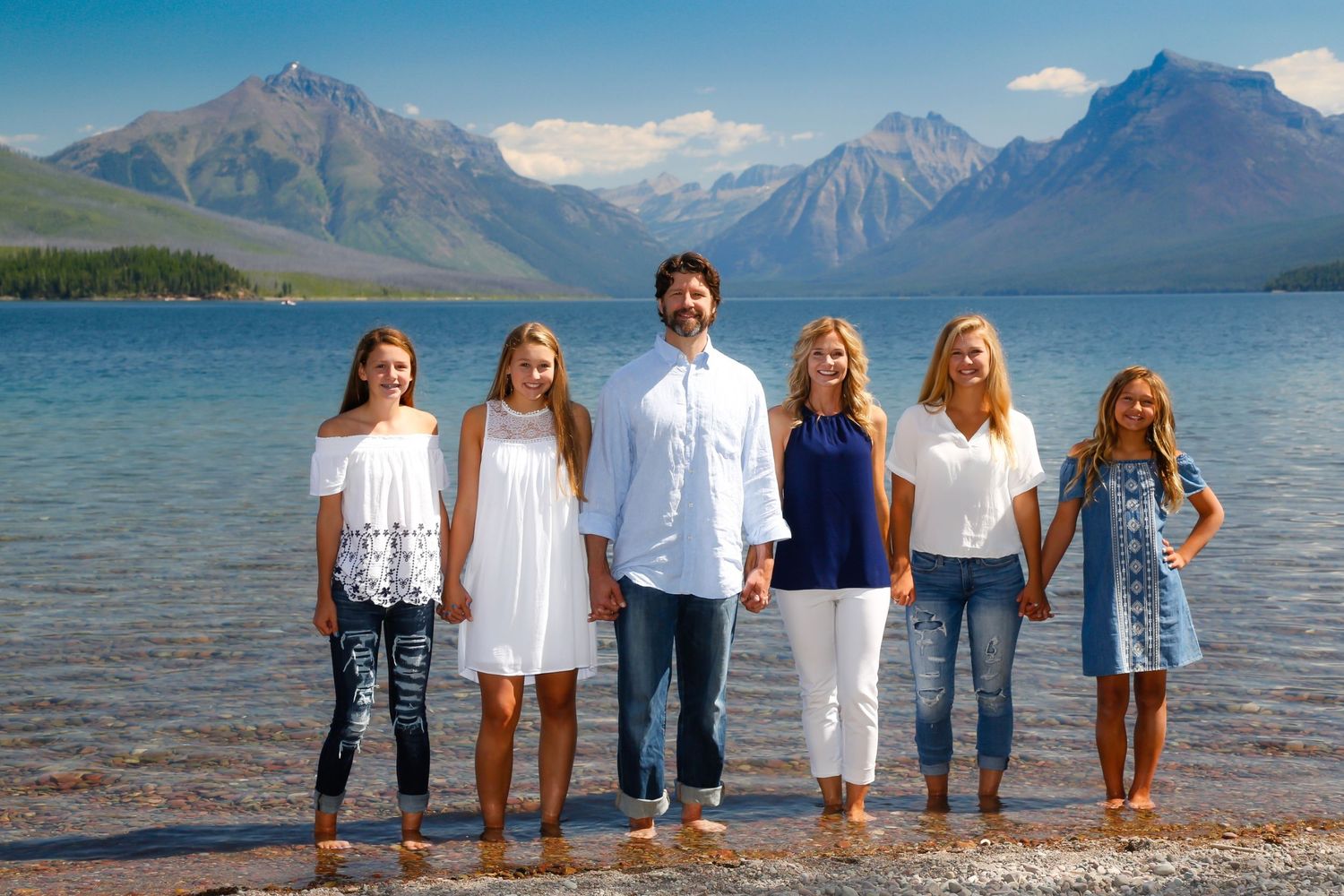 A family of six poses hand-in-hand on a lakeside beach with stunning mountain scenery behind them.