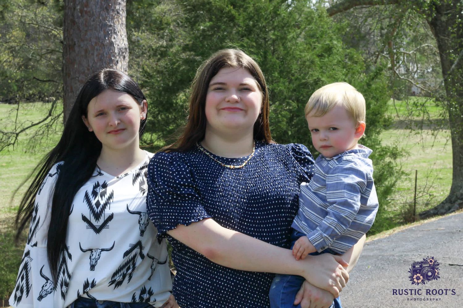 Two young women and a baby boy pose outdoors near trees at a Rustic Roots photography session.