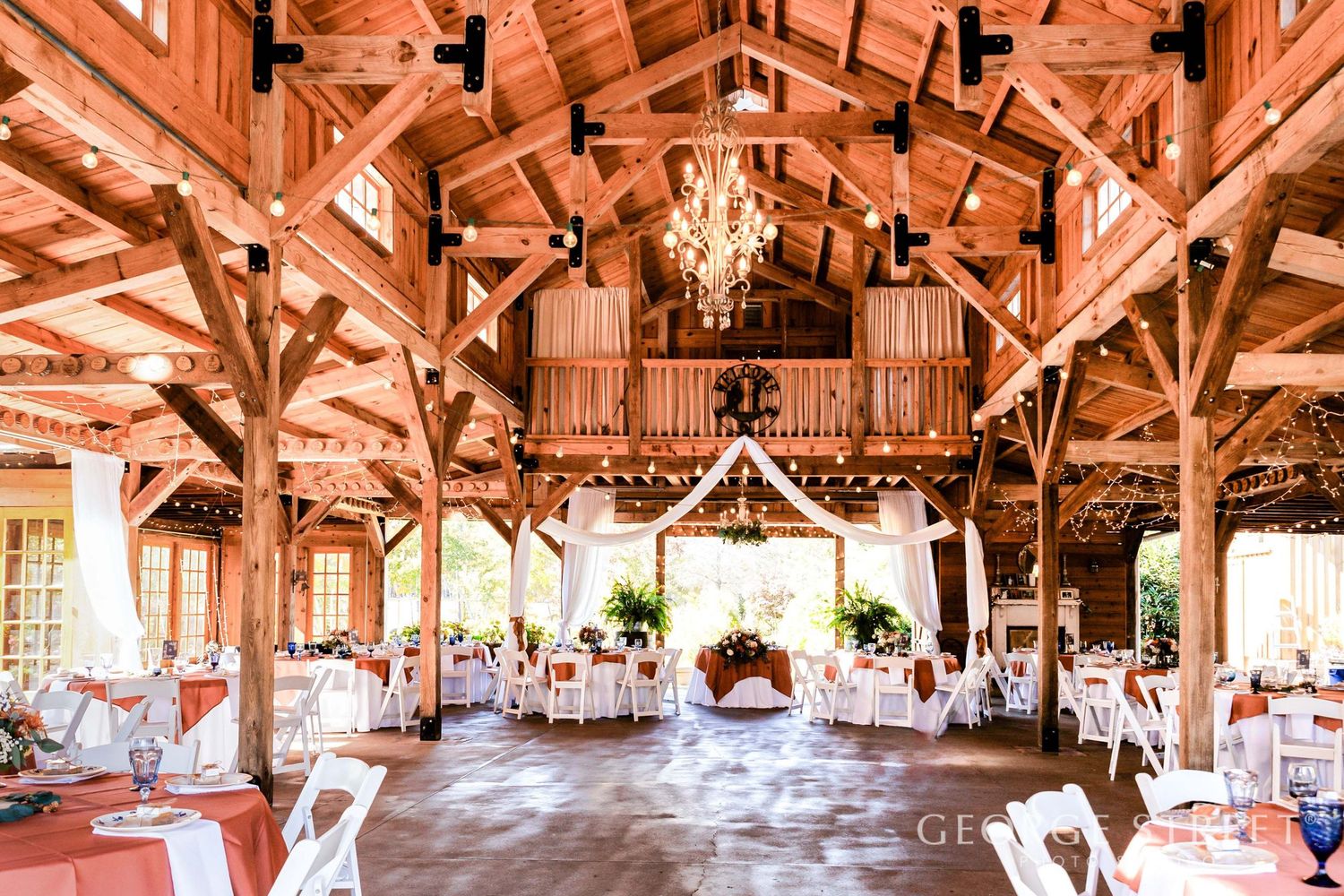 Rustic barn wedding venue with wooden beams, chandelier, white chairs, and elegantly decorated round tables for a reception.