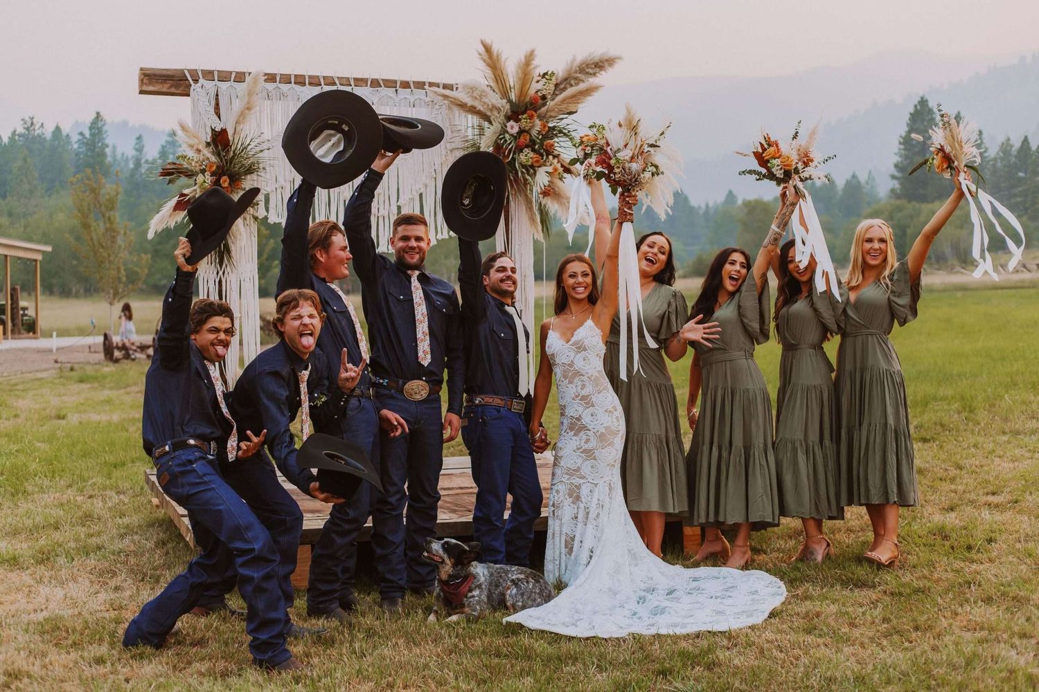 Wedding party celebrating outdoors in mountain setting, bride in white gown, bridesmaids in sage green dresses, groomsmen raising hats.